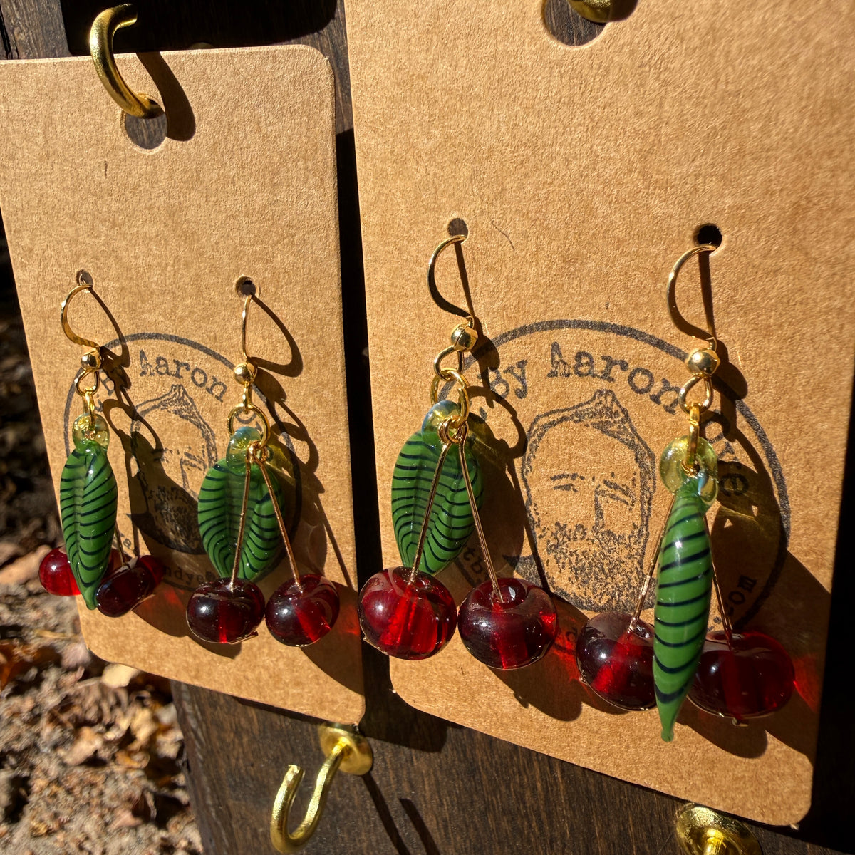 Two pairs of red glass cherry dangle earrings with gold hooks on kraft cards outdoors