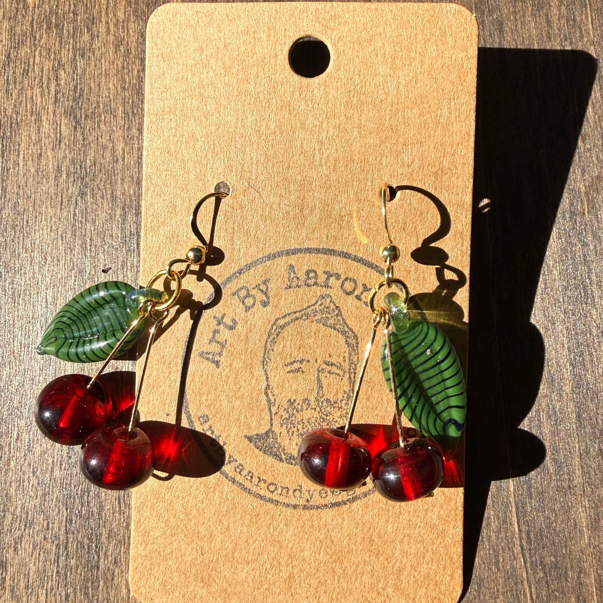 Red glass cherry earrings with striped green glass leaves on a kraft card against weathered wood