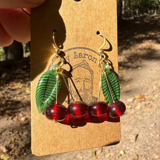 Hand blown glass cherry earrings with green leaves on an Art By Aaron Dye card along a sunny wooded trail