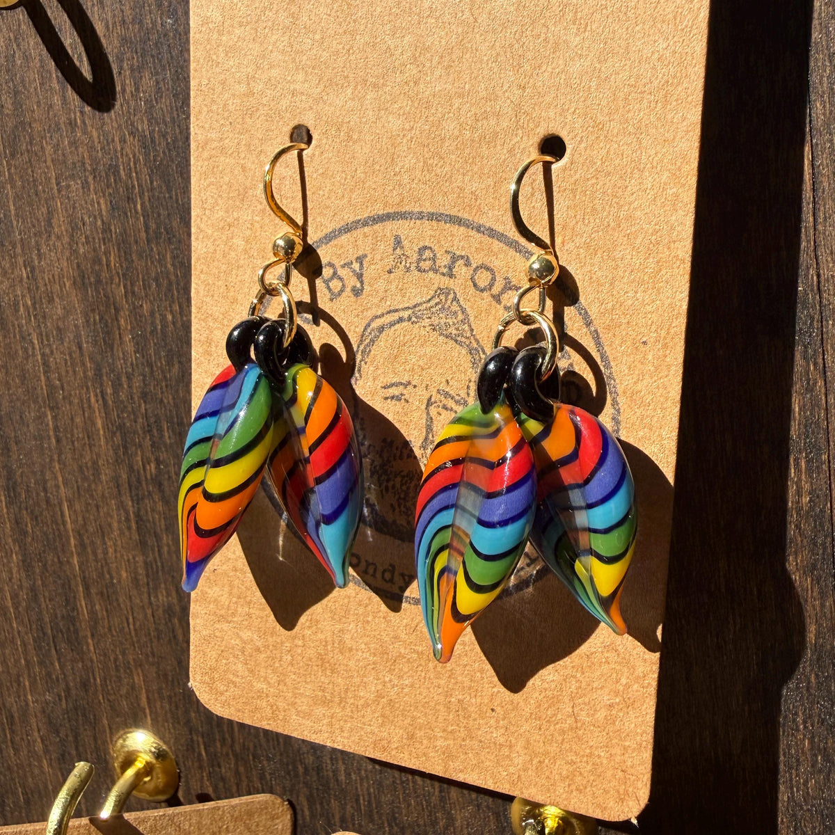 Rainbow striped hand blown glass double leaf earrings on gold hooks against a wood background